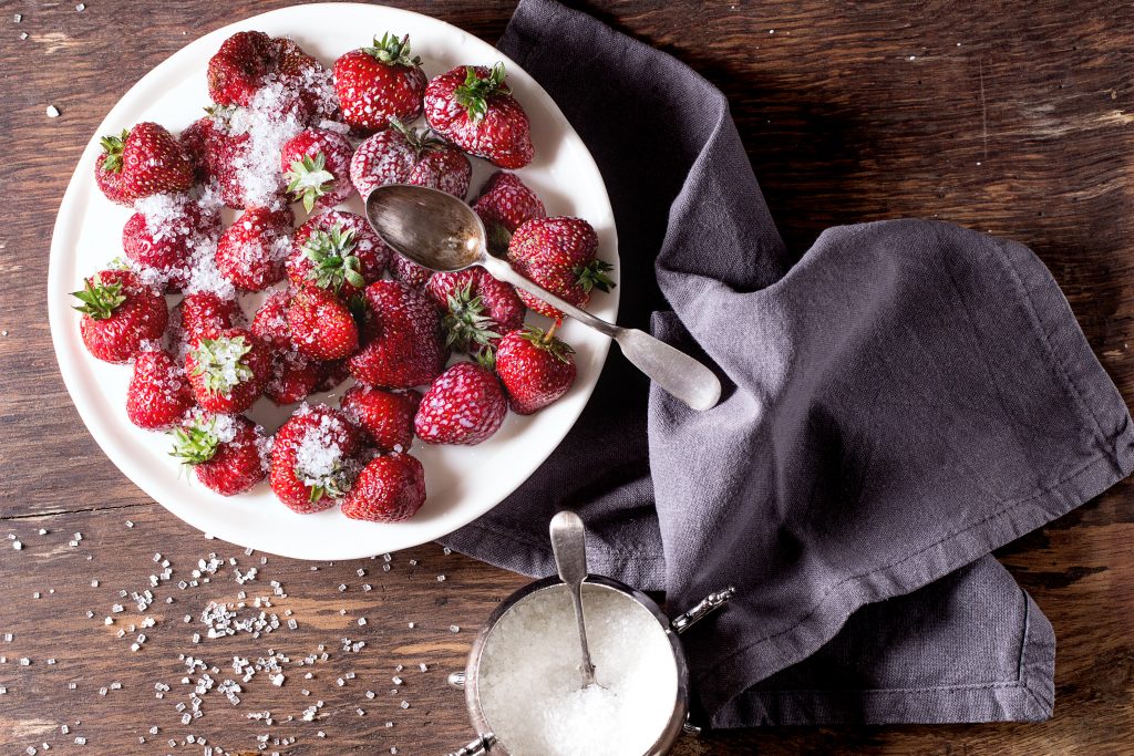 White plate with fresh ripe garden strawberries with sugar, sugar-bowl and spoons on wooden table. Dark rustic style. Top view